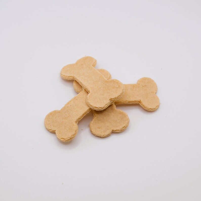May include: A pile of tan, bone-shaped dog treats on a white background. The treats have a slightly textured surface and are arranged in a casual, overlapping manner. The treats are a light brown color.