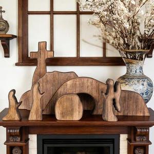 May include: Wooden Easter scene with a cross, figures, and a tomb. The figures are carved from wood and arranged on a dark wooden mantel. A vase with white flowers and a blue and white patterned vase are in the background.