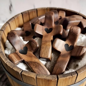 Puede incluir: Cruces de madera con un diseño de corazón negro se exhiben en un barril de madera. Las cruces están hechas de madera marrón claro con vetas visibles. El barril es de madera marrón claro con aros metálicos.