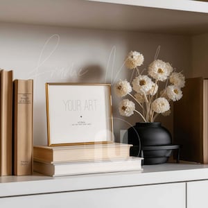 May include: A gold-framed picture with the text "YOUR ART" sits atop a stack of books. A black vase with white flowers is on a small black stand. Books with tan covers are on either side of the frame.