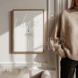 Könnte beinhalten: Ein gerahmtes Kunst-Mockup mit Holzrahmen, das den Text "YOUR ART VIDEO MOCKUP" anzeigt. Der Rahmen ist an einer weißen Wand montiert. Der Text "DIN A / Near 57" ist ebenfalls sichtbar. Eine Person trägt einen beigen Pullover.