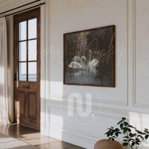 Può includere: Un'opera d'arte incorniciata con una scena serena di cigni bianchi che nuotano su uno specchio d'acqua scuro e riflettente. Il dipinto è incorniciato in una cornice di legno scuro e appeso a una parete bianca. La stanza ha una porta di legno e un pouf intrecciato.