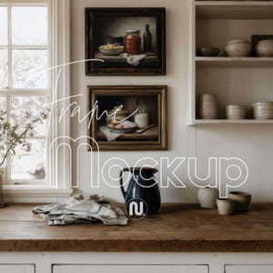 May include: A rustic kitchen scene featuring framed still-life paintings, a dark blue pitcher, and various ceramic bowls. A folded striped cloth rests on a wooden countertop. The word "Frame Mockup" is written in white.