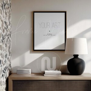 May include: A framed print with the text "YOUR ART" in a minimalist style. The frame is black with a gold border, and the print is displayed on a white wall. A black lamp with a white lampshade and a wooden cabinet are also visible.