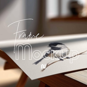 May include: A watercolor print of a small bird perched on a branch, with the text "Frame Mockup" overlaid in white. The print is displayed on a wooden table, with natural light illuminating the scene.
