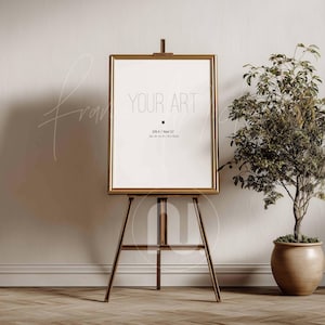 May include: A gold-framed art print with the text "YOUR ART" displayed on a wooden easel. The print is white with black text. A potted plant sits to the right of the easel. The room has a neutral color palette.