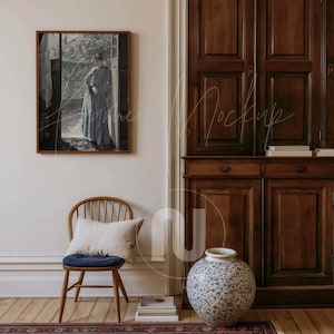 May include: A framed artwork depicts a woman in a long blue dress standing in a doorway. A wooden chair with a blue cushion and a decorative vase with a blue and white floral pattern are in the foreground. The room has wooden floors and a dark wood cabinet.