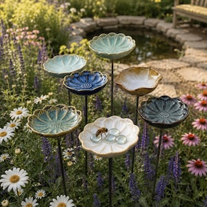 May include: A selection of ceramic flower-shaped bird baths in shades of blue, green, and white, set in a garden. Each bath is mounted on a black metal stake. A bee is seen on one of the baths, with a pond in the background.
