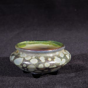 May include: A small, green and white ceramic pot with a speckled glaze. The pot has a rounded shape and three small feet.