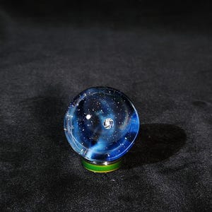 May include: A spherical glass paperweight featuring a deep blue and black nebula design with white speckles resembling stars and a central white spiral galaxy pattern. The orb rests on a small, circular, metallic green and gold base.