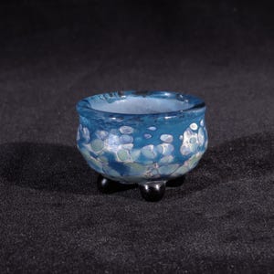 May include: A small, blue glass bowl with white and green flecks. The bowl has three black feet.
