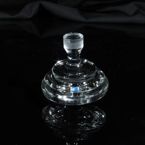 May include: A clear glass bottle stopper with a cylindrical top and a tiered, circular base. The stopper is made of clear glass and reflects light, creating a sparkling effect. The design is simple and elegant.
