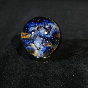 May include: A glass orb with a galaxy design. The orb features swirling blues, yellows, and whites, with tiny white speckles resembling stars. The orb sits on a dark, metallic base, creating a celestial effect.
