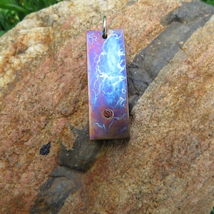 Puede incluir: Un colgante de metal azul y morado con un diseño de remolino blanco y un pequeño punto rojo en el centro. El colgante cuelga de una cadena plateada.