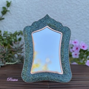 May include: Decorative mirror with a teal and copper frame. The frame has a textured, speckled design. The mirror is rectangular with a curved top. The mirror is sitting on a wooden surface.