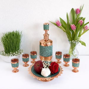 May include: A copper-colored decorative set featuring a tall bottle and small glasses with teal accents. The set is arranged with a red apple, rose, and garlic on a decorative plate, and a vase of pink tulips.