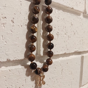 May include: A necklace made of large, round tiger's eye beads in brown and gold tones. It has gold-coloured metal accents and a gold letter "R" pendant. The necklace is displayed against a white brick wall.