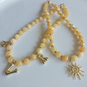 May include: Two necklaces with yellow and gold beads. One has gold letter charms spelling "CO" and "R". The other has a gold sun charm. The necklaces are on a white surface.