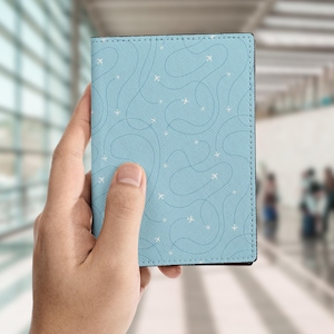 Op de afbeelding: Een lichtblauwe paspoorthouder met een patroon van witte vliegtuigen en gestippelde vliegroutes. De hoes wordt in een hand gehouden, met een vage achtergrond.