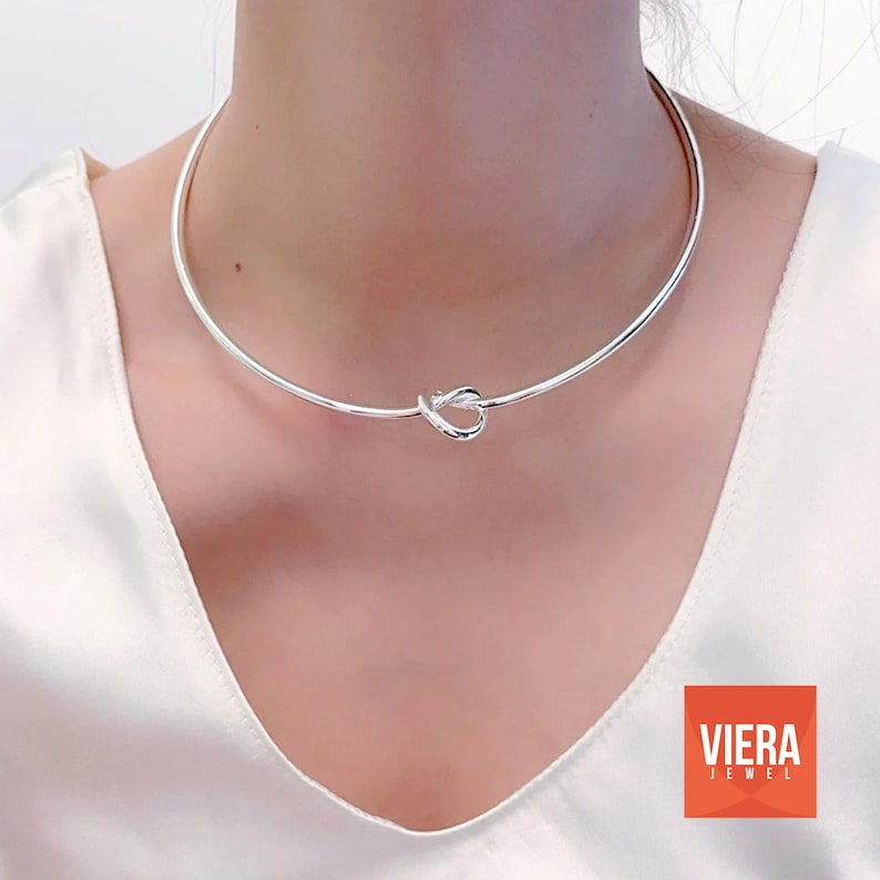 Może przedstawiać: Srebrny naszyjnik typu choker z węzłem. Naszyjnik, wykonany z błyszczącego metalu, jest noszony wok&oacute;ł szyi. To prosta, elegancka biżuteria. Widoczne jest r&oacute;wnież logo Viera Jewel.