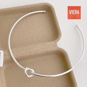 Może przedstawiać: Srebrny naszyjnik typu choker z węzłem pośrodku. Naszyjnik to prosty, elegancki element biżuterii. Nazwa marki "VIERA JEWEL" jest wyświetlana w czerwonym kwadracie w prawym g&oacute;rnym rogu.