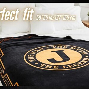 May include: A black blanket with a gold design and the text "THE MAN THE MYTH THE LEGEND" and a large letter "J". The blanket is on a bed with white pillows and a white sheet. The blanket measures 50 x 65 inches.