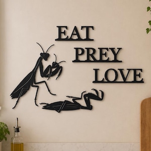May include: A black wall-mounted sign featuring the text EAT PREY LOVE arranged in three lines. To the left of the text, a black silhouette of a praying mantis is shown standing over another praying mantis lying on its back on the wall surface.