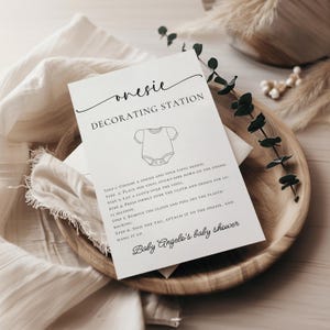May include: A white card with the text "onesie DECORATING STATION" and instructions for decorating a onesie. The card is on a wooden tray with a white cloth and decorative greenery. The text "Baby Angelo's baby shower" is also visible.