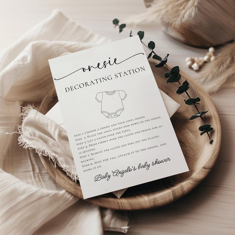May include: A white card with the text "onesie DECORATING STATION" and instructions for decorating a onesie. The card is on a wooden tray with a white cloth and decorative greenery. The text "Baby Angelo's baby shower" is also visible.