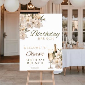 Puede incluir: Un letrero blanco sobre un caballete de madera con las palabras "Birthday Brunch" y "Welcome to Olivia's Birthday Brunch" en una elegante escritura. El letrero está decorado con detalles florales y una botella y copa de champán.