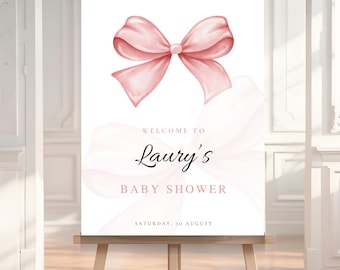 May include: A baby shower welcome sign on an easel. The sign features a watercolor illustration of a pink bow at the top. The text reads "Welcome to Laury's Baby Shower" and "Saturday, 10 August".
