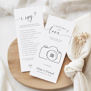 May include: Two white wedding cards with black text. One card says "i spy" with a list of photo prompts. The other says "Capture the love" with a camera graphic and a QR code. The cards are on a wooden tray.