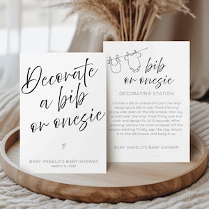 Op de afbeelding: Twee witte kaarten met zwarte tekst op een houten dienblad. Eén kaart leest "Versier een slabbetje of onesie" met een hartje en babyshower details. De andere kaart zegt "slabbetje of onesie decoratiestation" met instructies.