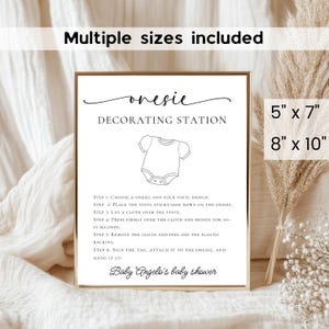 May include: A framed print with the text "onesie DECORATING STATION" and instructions for decorating baby onesies. The print includes the text "Baby Angelo's baby shower" and is available in 5" x 7" and 8" x 10" sizes.