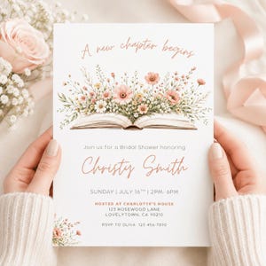 May include: A white bridal shower invitation card featuring an open book illustration with a bouquet of pink and white wildflowers emerging from the pages. The text reads A new chapter begins, Join us for a Bridal Shower honoring Christy Smith.