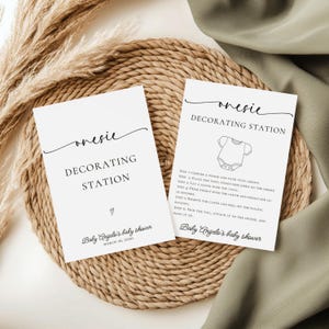 May include: Two white cards with the text "onesie DECORATING STATION" are displayed on a woven mat. One card includes instructions and a onesie graphic. The cards are for Baby Angelo's baby shower, March 16, 2030.
