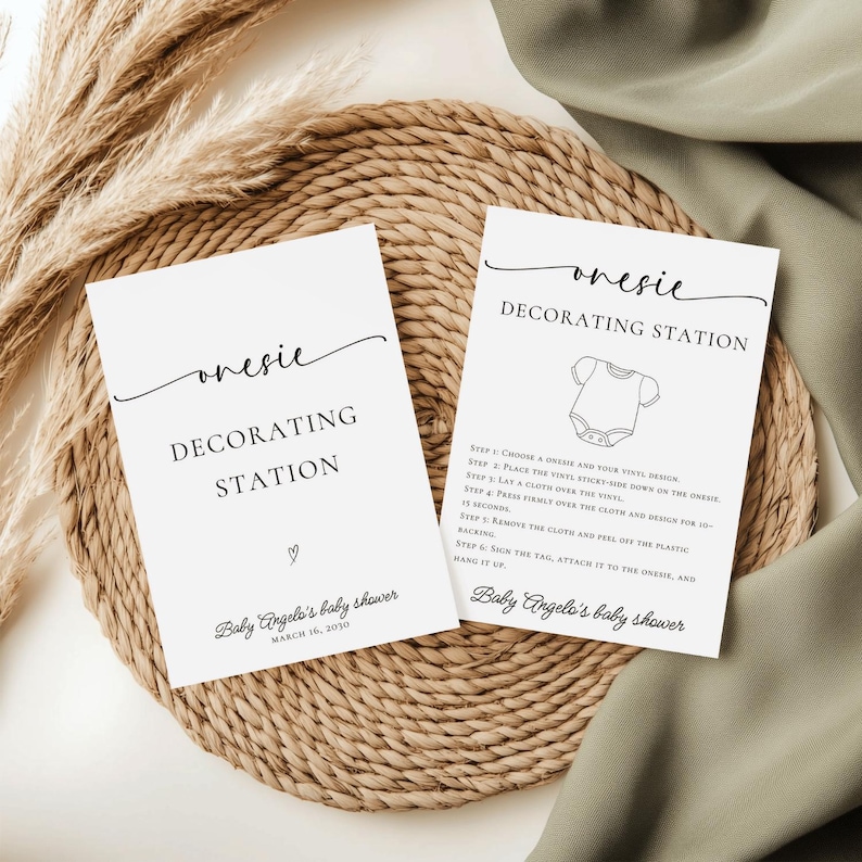 May include: Two white cards with the text "onesie DECORATING STATION" are displayed on a woven mat. One card includes instructions and a onesie graphic. The cards are for Baby Angelo's baby shower, March 16, 2030.