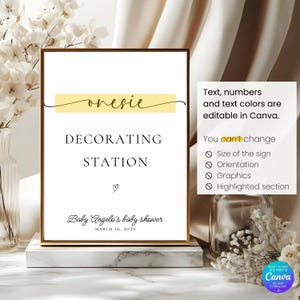 May include: A framed sign with the words "onesie DECORATING STATION" in black font, with a yellow banner above. Below, it reads "Baby Angelo's baby shower MARCH 16, 2030". The sign is on a marble base.