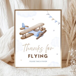 May include: A framed print with a watercolor illustration of a biplane, clouds, and stars. The text reads "Thanks for FLYING, PLEASE TAKE A FAVOR." The artwork has a gold frame and is set against a neutral background.