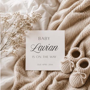 Könnte beinhalten: Eine quadratische weiße Ankündigungskarte mit schwarzem Text BABY Lavian IS ON THE WAY DUE APRIL 20XX, begleitet von einem Paar kleiner beiger gestrickter Babyschuhe und einem Zweig getrockneter weißer Schleierkrautblüten auf einem strukturierten Stoff.