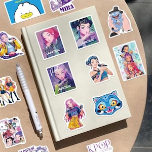 May include: A collection of colorful stickers featuring K-pop singers and cartoon characters. The stickers are placed on a white notebook and a wooden surface. The stickers include illustrations of women with microphones, a blue cat, and the text "HUNTR/X".
