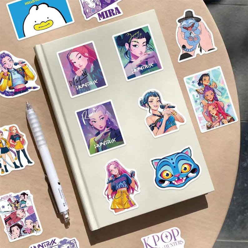 May include: A collection of colorful stickers featuring K-pop singers and cartoon characters. The stickers are placed on a white notebook and a wooden surface. The stickers include illustrations of women with microphones, a blue cat, and the text "HUNTR/X".