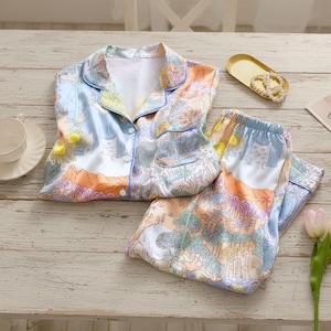 Puede incluir: Un conjunto de pijama de dos piezas con un estampado de paisaje en tonos azules, naranjas y verdes. La parte superior tiene una botonadura y un cuello con solapas. Los pantalones tienen una cintura elástica. El conjunto es de material sedoso.