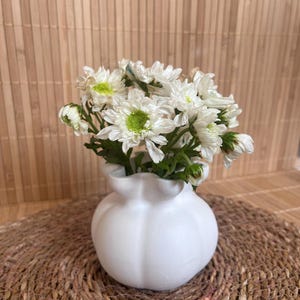 May include: A small white vase with a ruffled top holds a bouquet of white flowers with green centers. The vase sits on a round, woven mat. The background is a light brown bamboo mat.