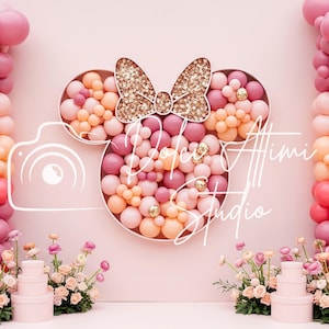 May include: A pink and peach balloon arch with a Minnie Mouse head design, featuring a gold glitter bow. The backdrop includes floral arrangements and stacked gift boxes, creating a festive party scene.