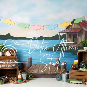 May include: A photography backdrop featuring a lake scene with a wooden dock, a rustic cabin, and a garland of colorful fish. Wooden crates hold fishing-themed props, including a fishing lure and buoys. The text "Gone Fishin' Bait & Tackle" is visible.