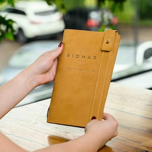 May include: A tan leather portfolio with the text "HOTEL RIOMAR IBIZA TRIBUTE PORTFOLIO" embossed on the front. The portfolio is held open, revealing the interior. A buttoned flap secures the portfolio.