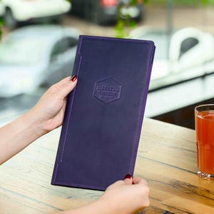 以下が含まれることがあります： 紫色の革製メニューを人が持っています。「Reserva Restaurante」のエンボス加工が六角形に施されています。オレンジジュースのグラスが、メニューの隣の木製のテーブルに置かれています。