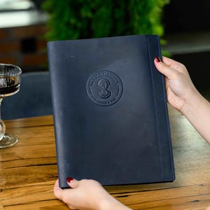 May include: A navy blue leather menu cover with embossed logo and text. The menu is held by a person with red nail polish. A latte and a drink in a stemmed glass are on a wooden table.