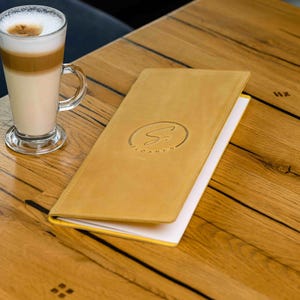 May include: A latte in a clear glass with a handle sits on a wooden table next to a yellow leather menu. The menu has a circular logo with the word "SILVANO" embossed on the cover. The table has a natural wood grain.